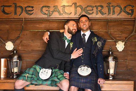 Two grooms wearing Highlandwear sitting on wooden bench with wooden sign saying 'The Gathering'