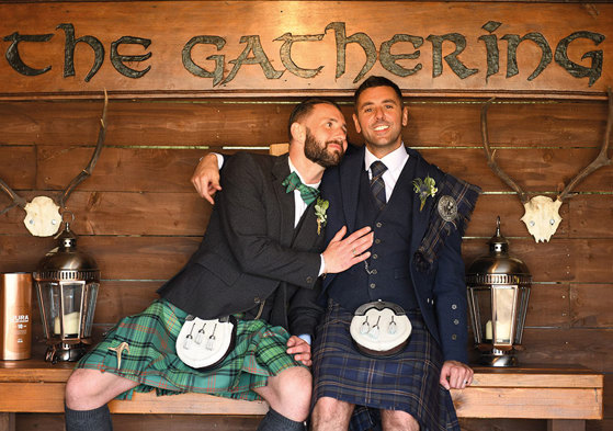 Two grooms wearing Highlandwear sitting on wooden bench with wooden sign saying 'The Gathering'