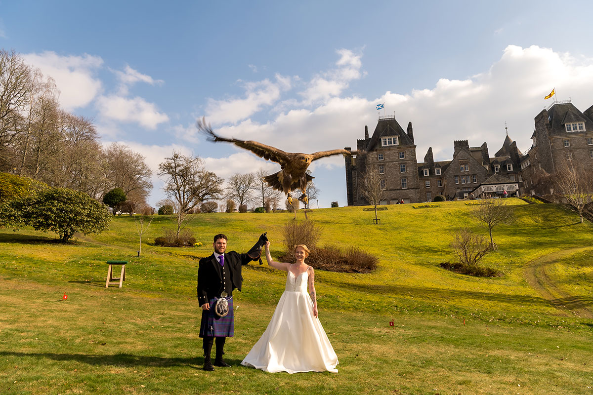 Bride and groom posing on the lawn at Atholl Palace Hotel as a bird of prey flies overhead, with the castle hotel and gardens behind them