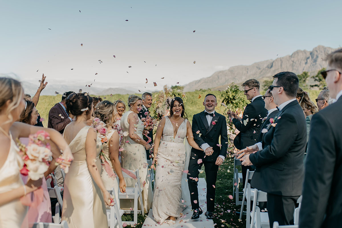 Newlyweds smiling as guests throw rose petal confetti during their outdoor South Africa wedding ceremony at Boschendal Wine Estate