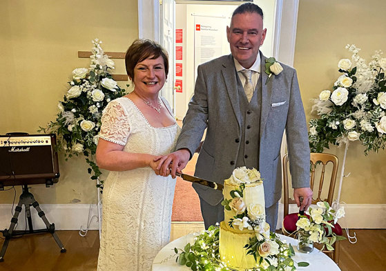 A bride and groom cut their two tier wedding cake which is covered in greenery and white flowers 