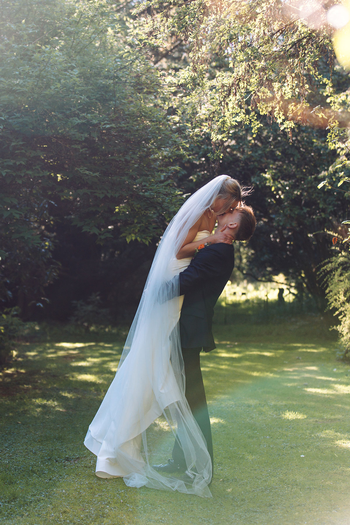 Bride lifted by groom during outdoor wedding portraits in sunlit garden setting