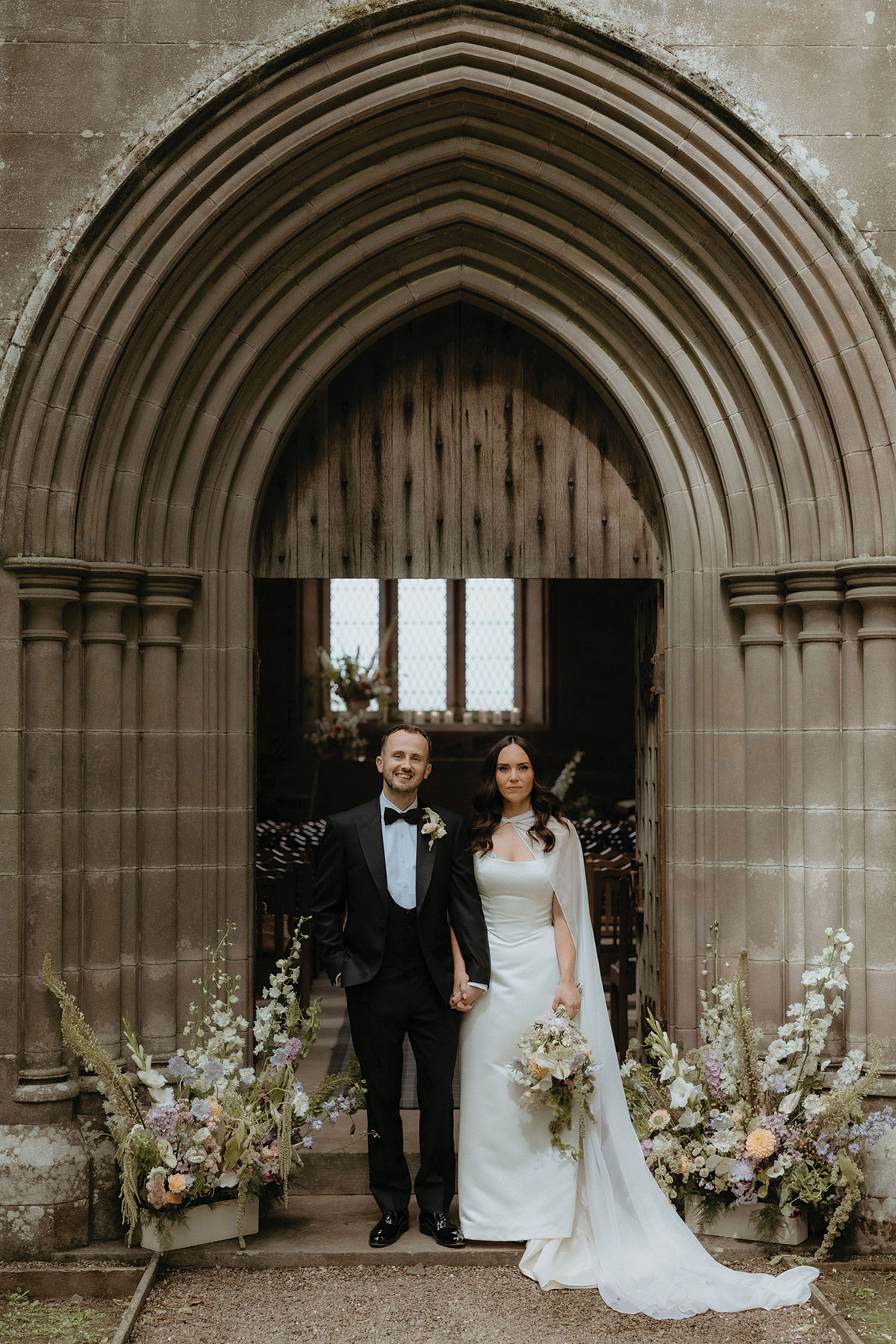 Couple standing at stone church doorway with pastel floral ceremony arrangements
