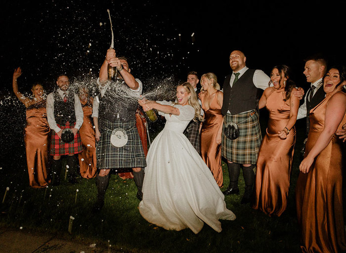 a bride and groom doing a champagne spray in the dark with men in kilts and women in rust coloured bridesmaid dresses cheering in the background 