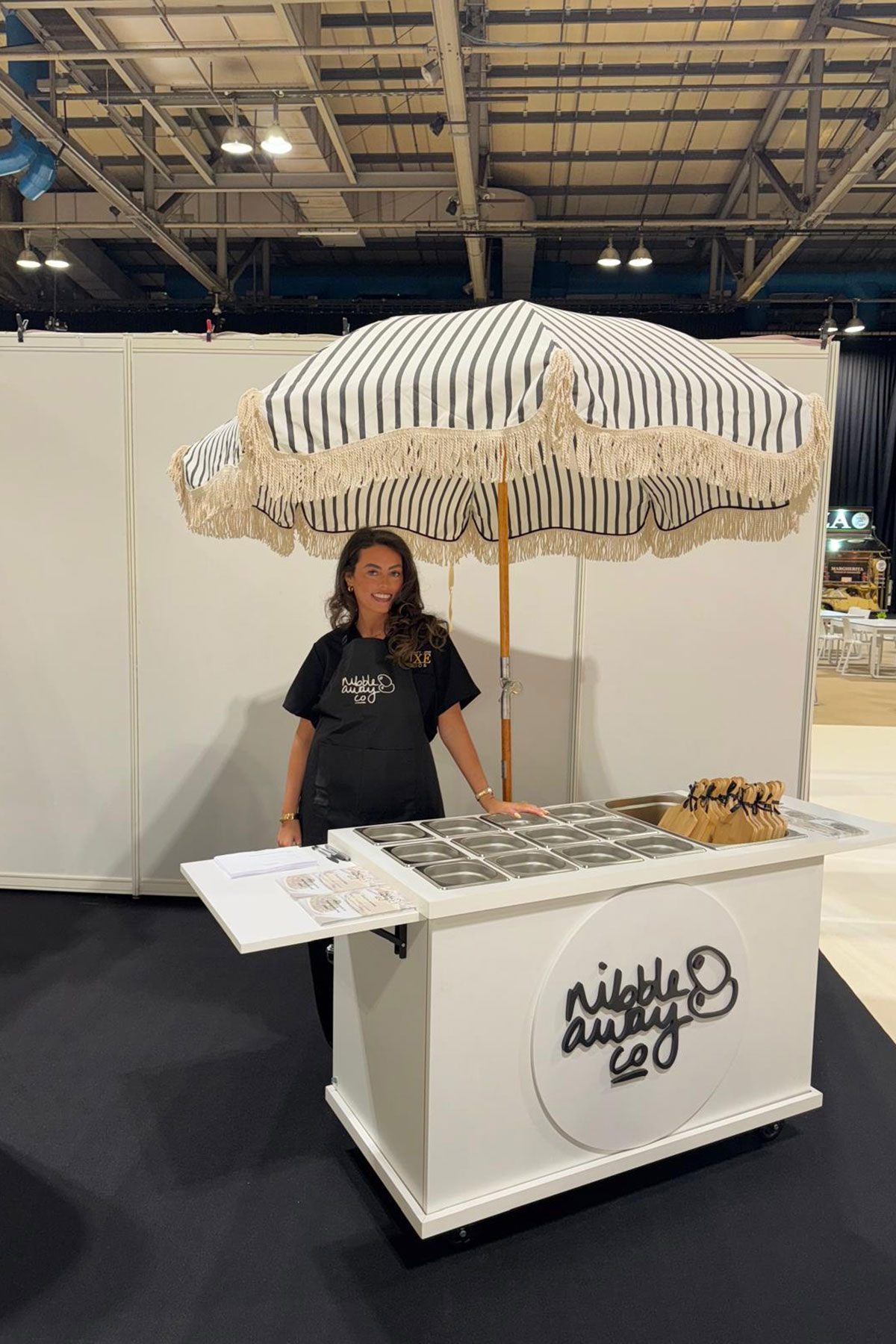 A woman wearing a Nibble Away apron stands behind a white mobile grazing cart with a striped parasol at an indoor event.