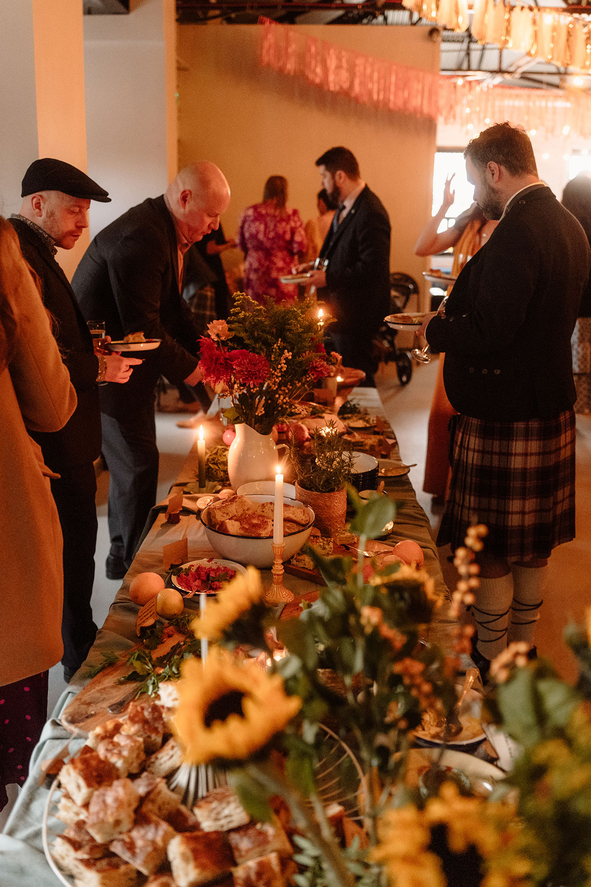 Guests gather around a beautifully styled sharing feast table with candles, seasonal flowers and rustic food displays during a wedding reception