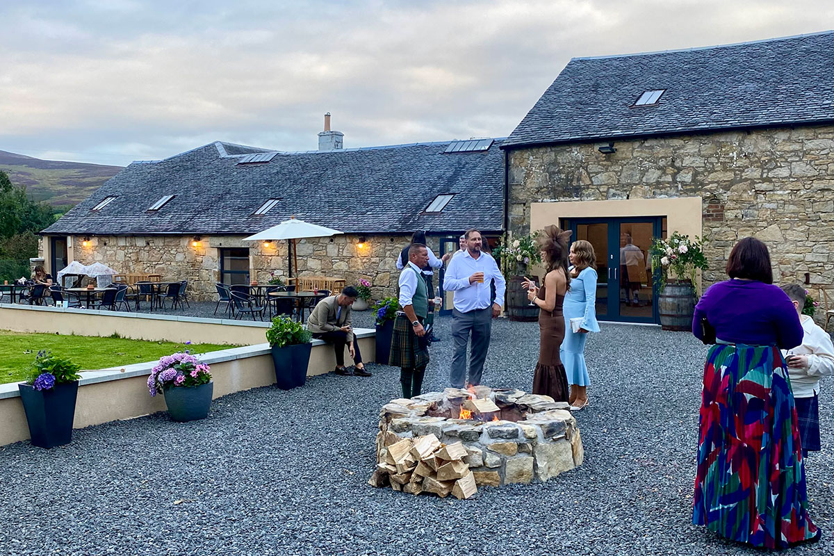 Evening drinks reception at Blackshaw Barns with guests chatting around a stone firepit on the courtyard terrace beside the converted barns.