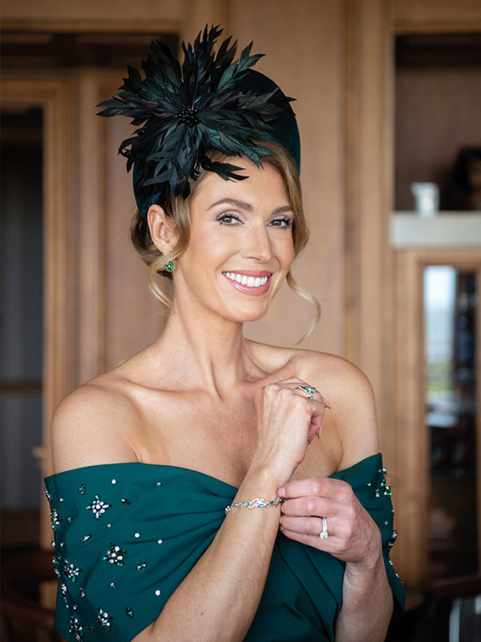 A woman wearing a dark green dress with sparkly beading and a dark green fascinator smiles at the camera as she adjusts her bracelet