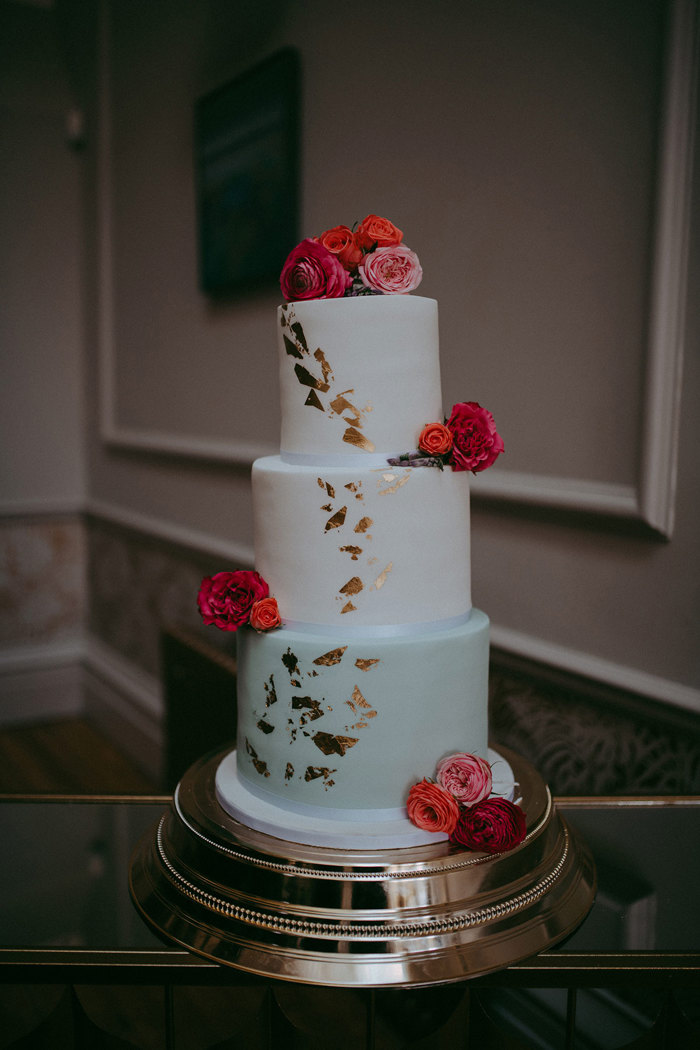 a three tier white and pale green wedding cake with gold leaf accent and pink and red flower decoration sitting on a chunky silver platter