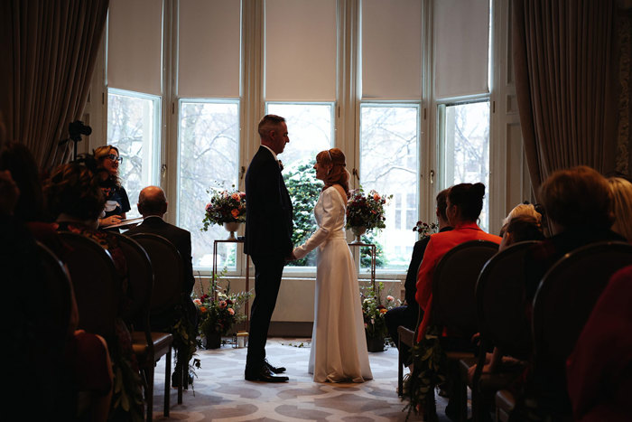 a bride and groom hold hands in front of a window with guests sitting either side of the aisle with their back to the camera 
