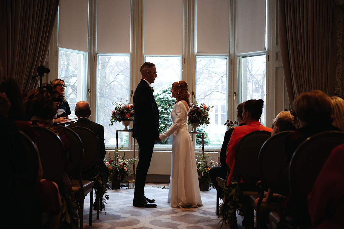 a bride and groom hold hands in front of a window with guests sitting either side of the aisle with their back to the camera