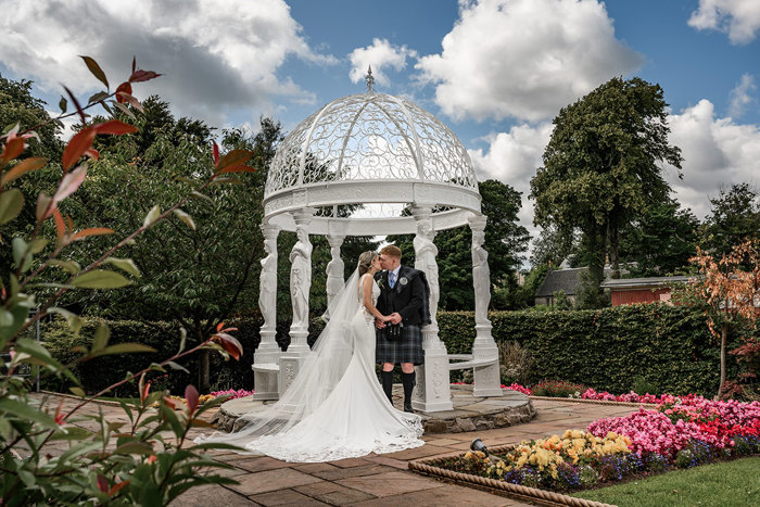 bride and groom stood outside kissing in garden alongside large dome marble sculpture