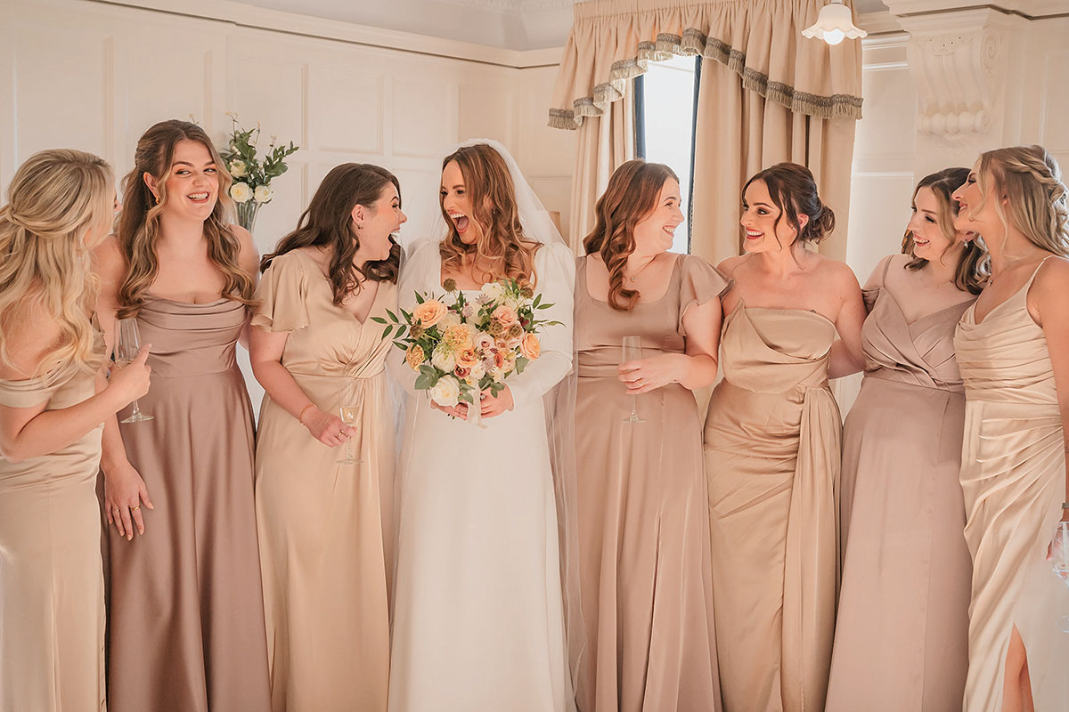 Bride holding a bouquet and laughing with bridesmaids wearing satin dresses in soft neutral tones