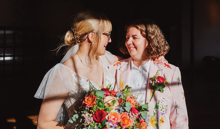 Brides smiling at each other in warm sunlight, holding a bright, colourful bouquet.