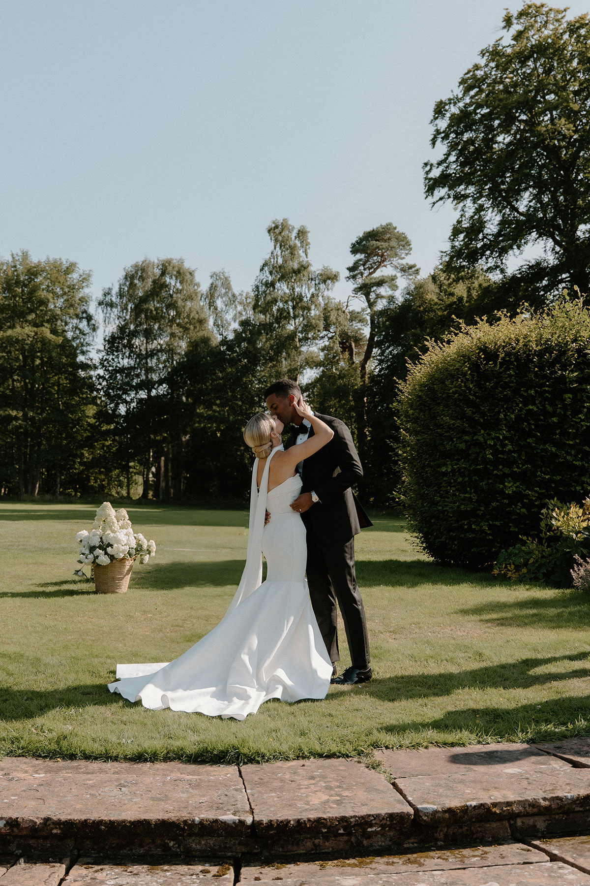Newlywed couple kissing outdoors after their garden wedding ceremony surrounded by greenery and white floral arrangements