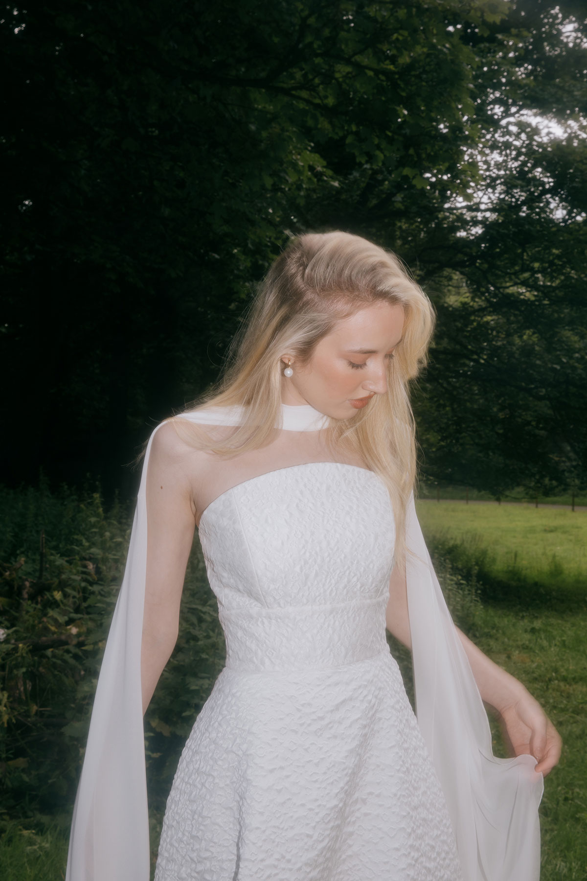 A bride in a textured strapless white gown looks down while standing in a grassy countryside area with trees in the background.