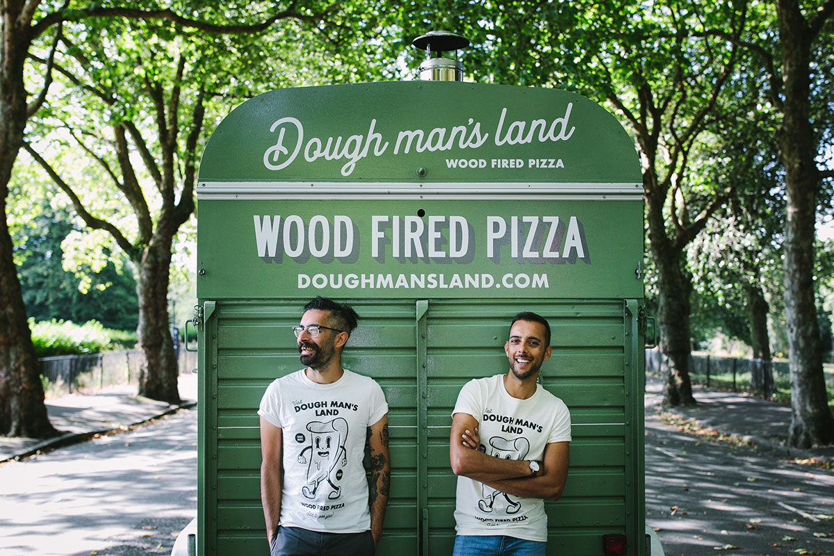 Two Dough Man’s Land team members wearing branded T-shirts standing in front of their wood-fired pizza trailer in a tree-lined park