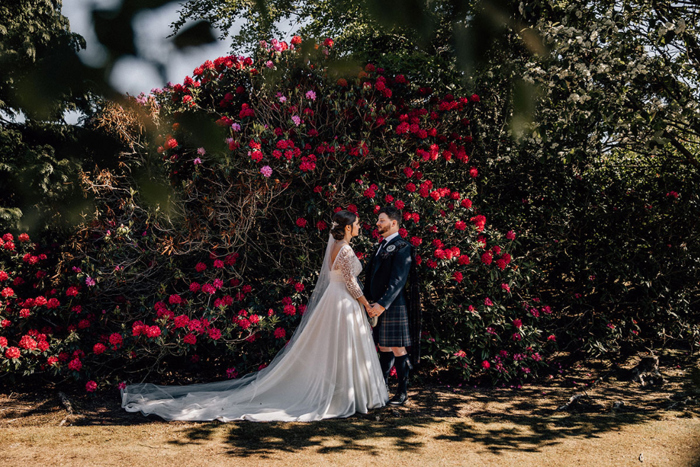 Bride and groom hold hands outside in front of bush with red flowers on it