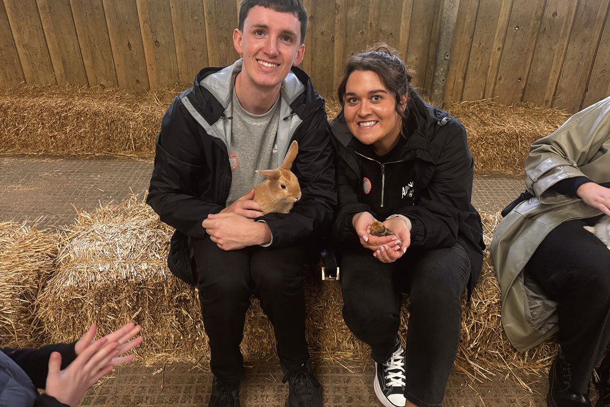 two people sitting on a bale of hay, one holding a small rabbit, the other a small chick