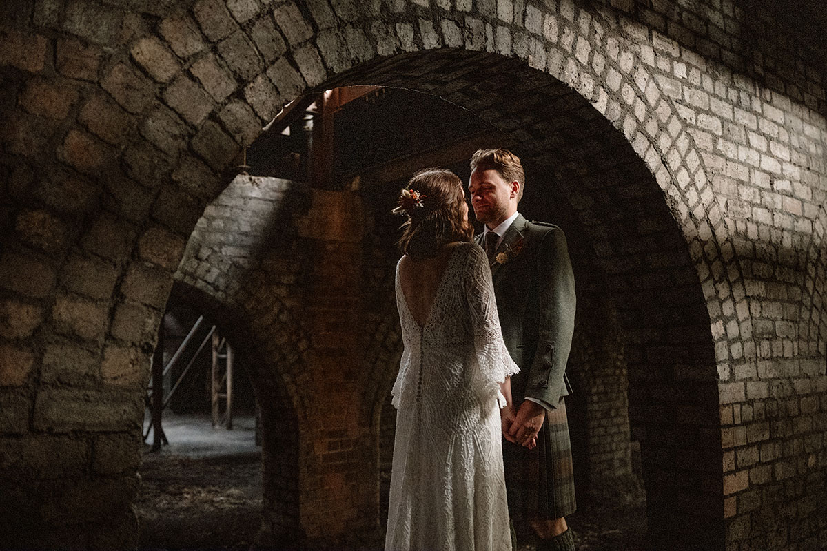 Bride and groom stand hand in hand beneath historic brick arches inside an industrial heritage venue