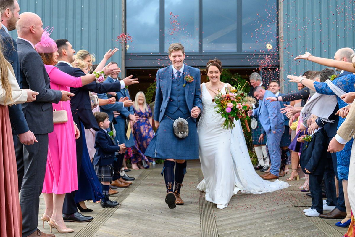 A bride wearing a long white dress and veil, and a groom wearing a blue kilt walk out of a building holding hands while their guests throw confetti over them