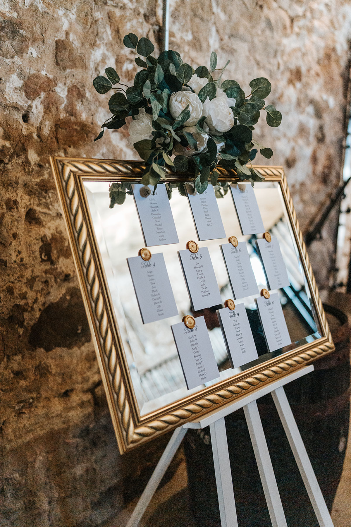 Gold-framed wedding seating plan mirror with white roses and eucalyptus on a white easel