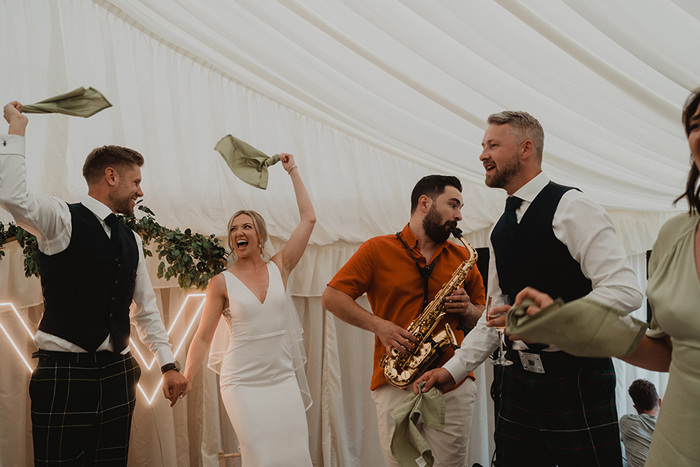A Bride And Groom Swinging Napkins Beside A Person Playing A Saxophone