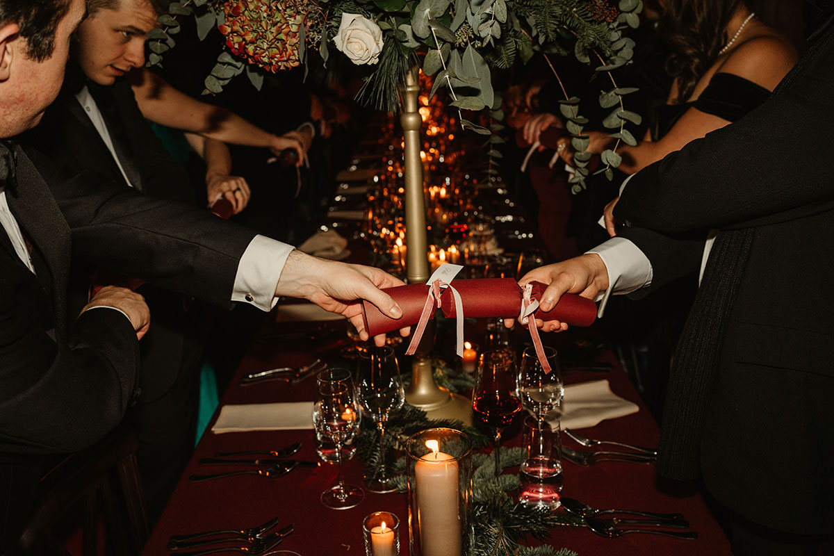 Guests reaching across the table to pull red Christmas crackers above a candle-lit dinner setting.