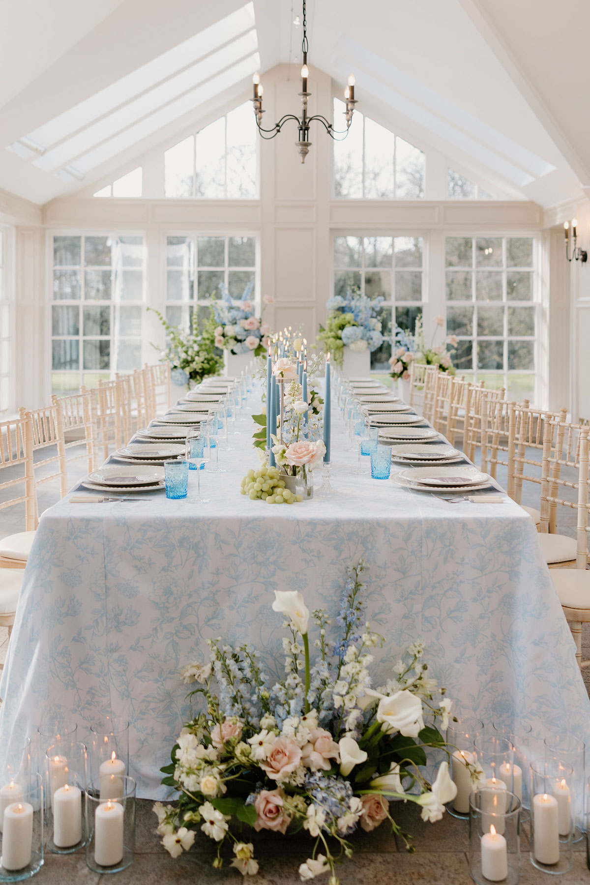 Reception table with blue floral table linen, candles and floral arrangements in a bright conservatory