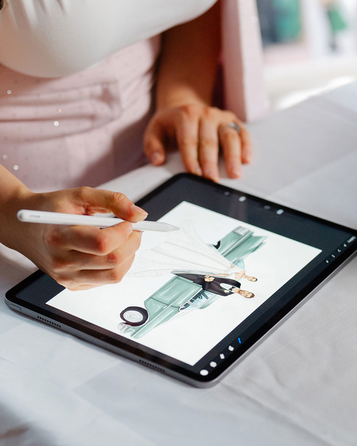 a hand holding a tech pen over an ipad, drawing a bride and groom standing in front of a car