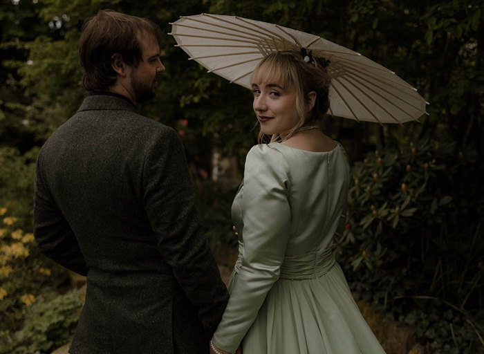 a bride wearing a light green dress and carrying a parasol walks with a groom wearing a tweed suit. There are bushes with flowers in the background