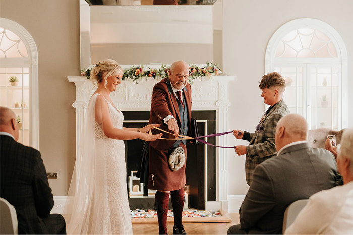 A man in a brown kilt conducts a handfasting ceremony with two pieces of fabric between two brides
