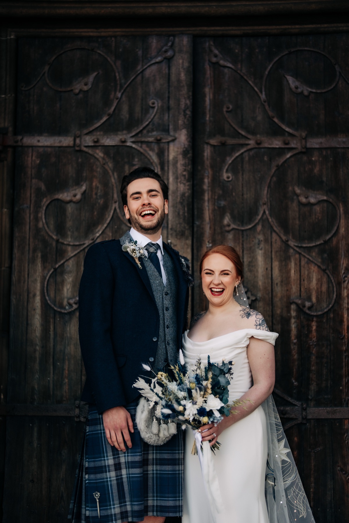 Newlyweds Robyn and James laugh together outside Òran Mór's wooden entrance doors, holding a blue-and-white bridal bouquet.