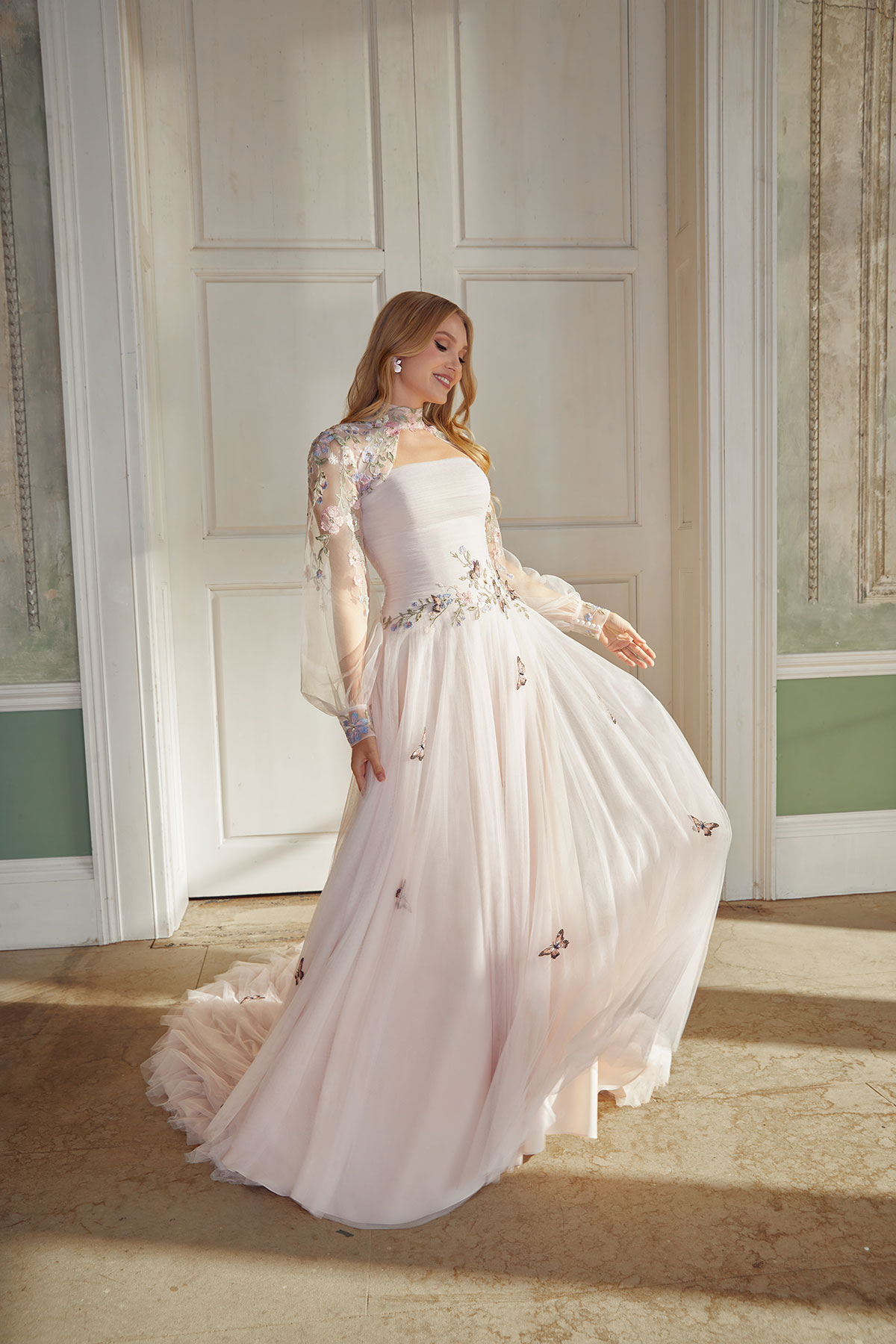 Bride wearing a blush-toned wedding dress with sheer embroidered sleeves, soft floral appliqué and a floaty skirt, photographed inside an elegant historic room