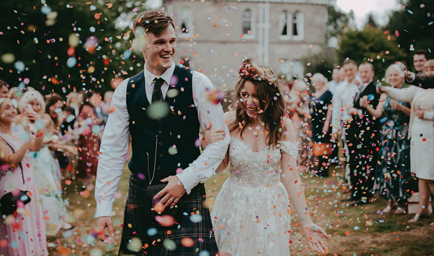 a smiling bride and groom walking arm in arm in front of an elegant building while being showered in colourful paper confetti