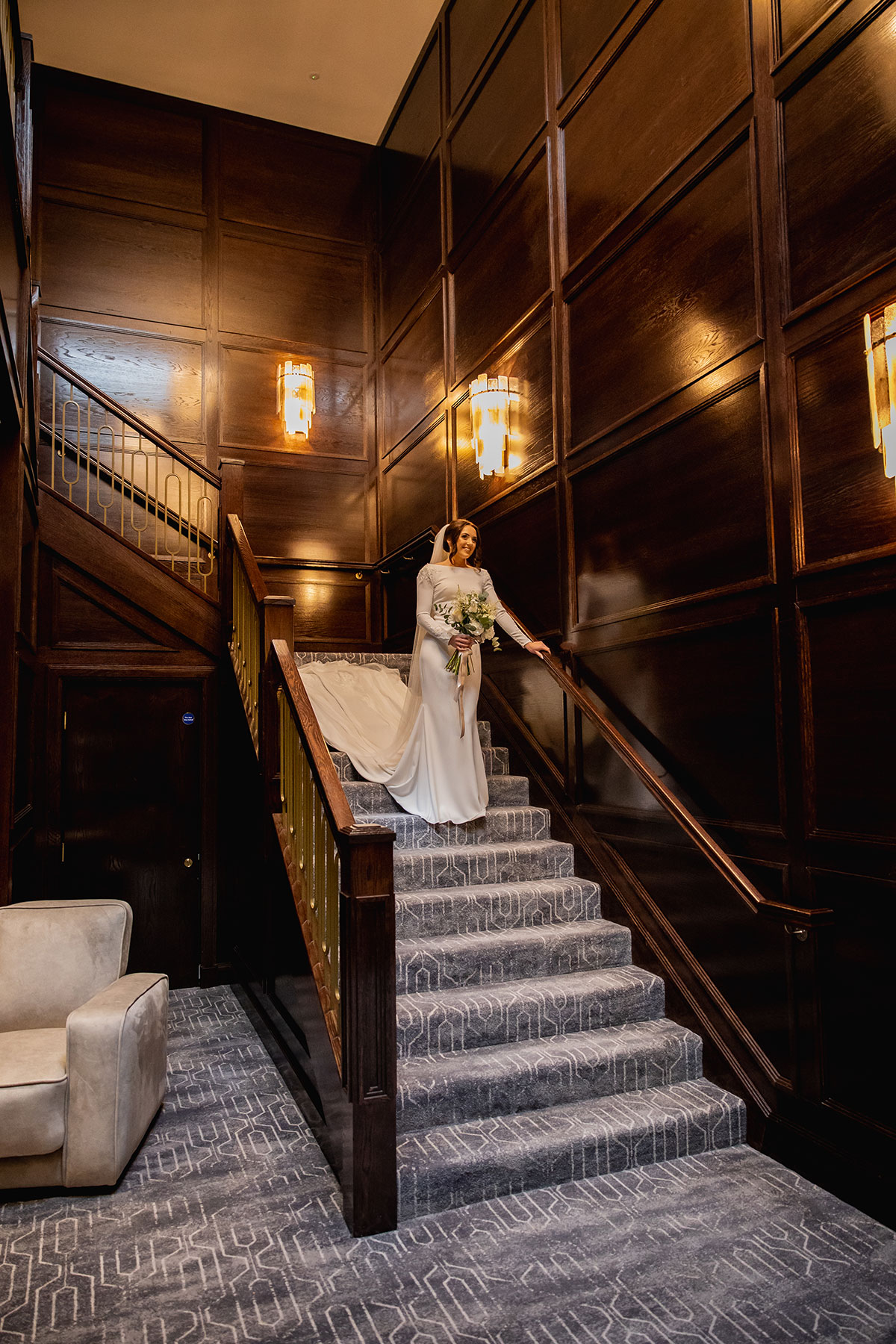 Bride in elegant white dress walking down a wooden staircase with golden lighting
