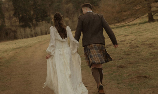 bride in trailing satin corset gown and groom in kilt outfit hold hands, running along a dirt path, surrounded by grass and a forrest of trees