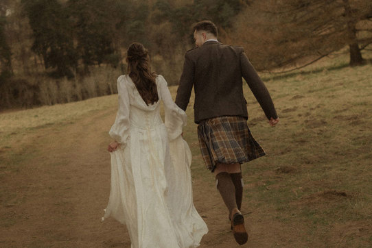 bride in trailing satin corset gown and groom in kilt outfit hold hands, running along a dirt path, surrounded by grass and a forrest of trees