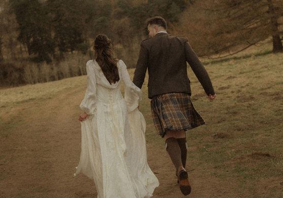 bride in trailing satin corset gown and groom in kilt outfit hold hands, running along a dirt path, surrounded by grass and a forrest of trees
