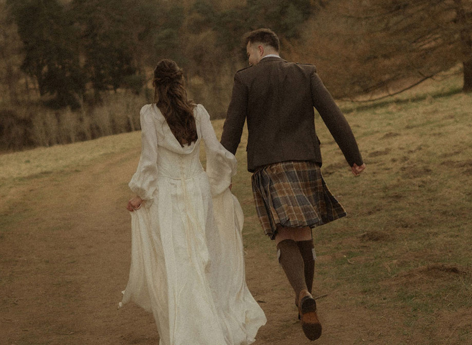 bride in trailing satin corset gown and groom in kilt outfit hold hands, running along a dirt path, surrounded by grass and a forrest of trees