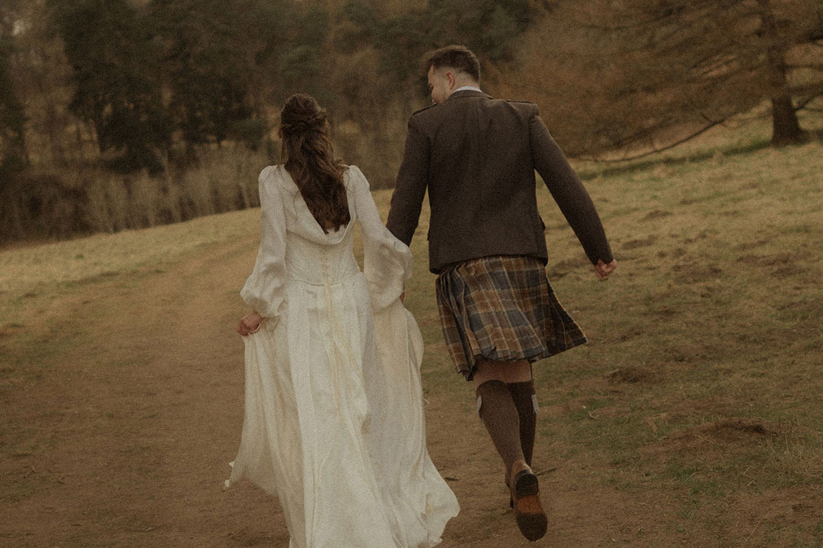 bride in trailing satin corset gown and groom in kilt outfit hold hands, running along a dirt path, surrounded by grass and a forrest of trees