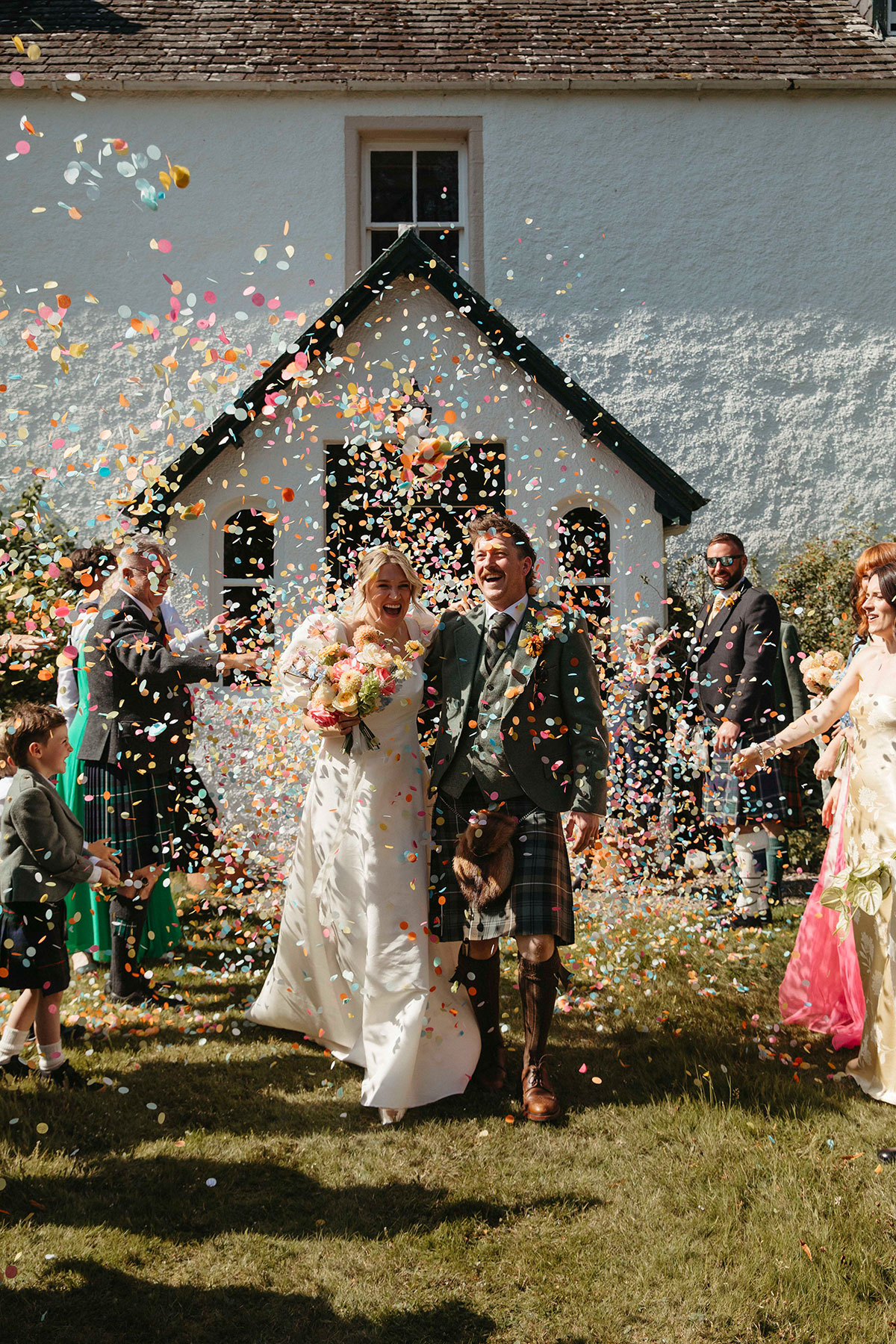 Bride and groom walk through colourful confetti outside bride’s family home during Cairngorms farmhouse wedding in Nethy Bridge