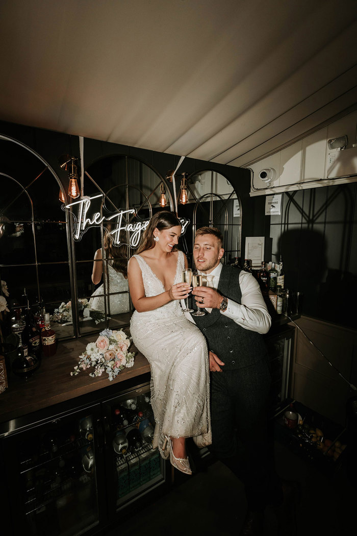 Alex and Matt Fagerson posing for a photo with champagne in the bar area at Dunglass Estate. There is a neon sign reading 'The Fagersons' behind them