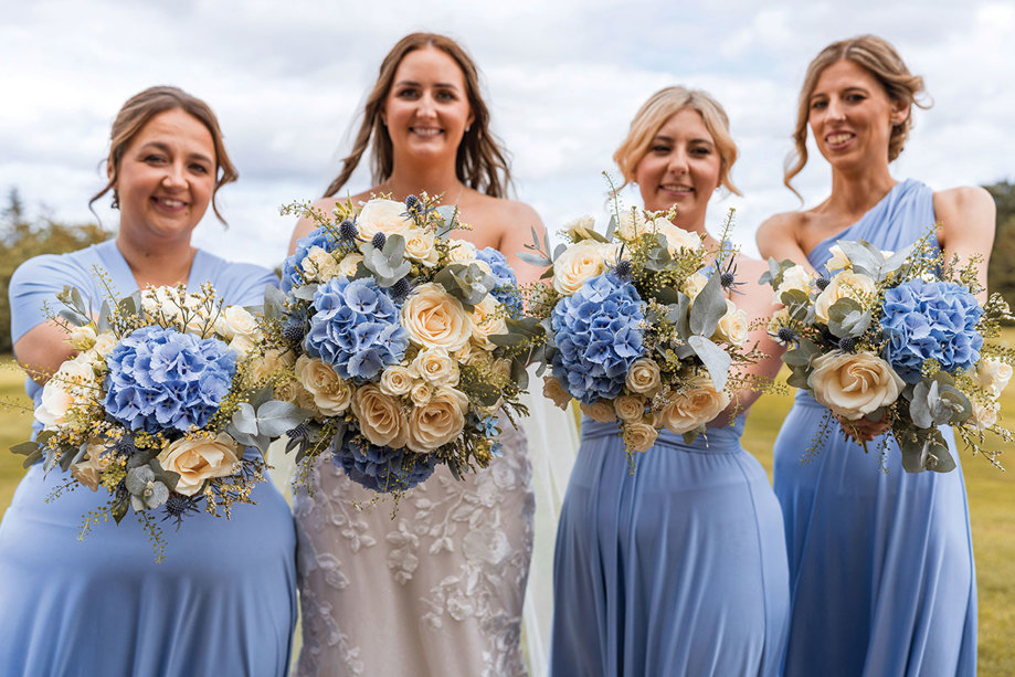 four women, consisting of three bridesmaids and one bride, hold their blue and white bouquets out towards the camera
