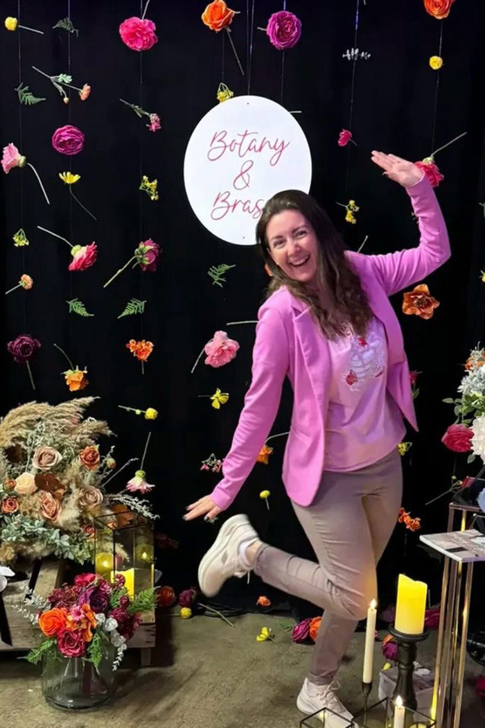 a brunette woman wearing a pink jacket posing in front of a colourful floral wall against a black background