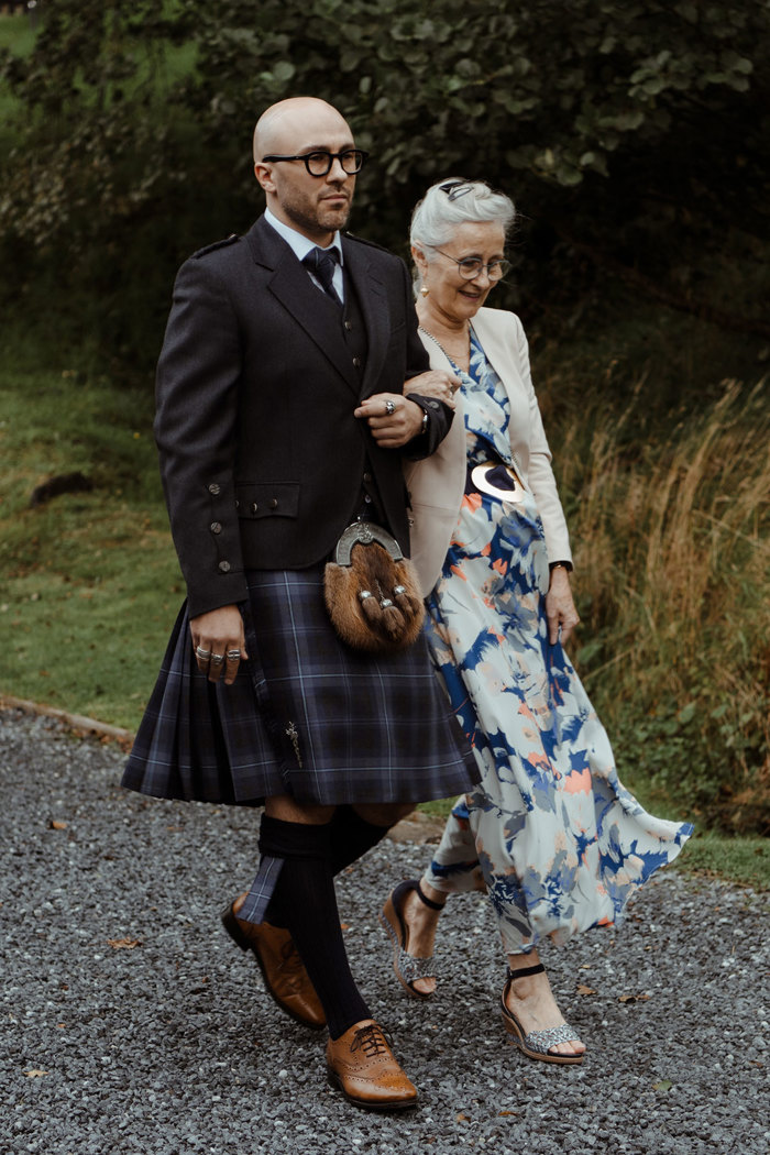 groom in blue, grey and white tartan kilt outfit with brown brogues and a brown sporran, holds on to the arm of an older white haired woman in a dark blue floral dress and cream jacket as they walk across a gravel path