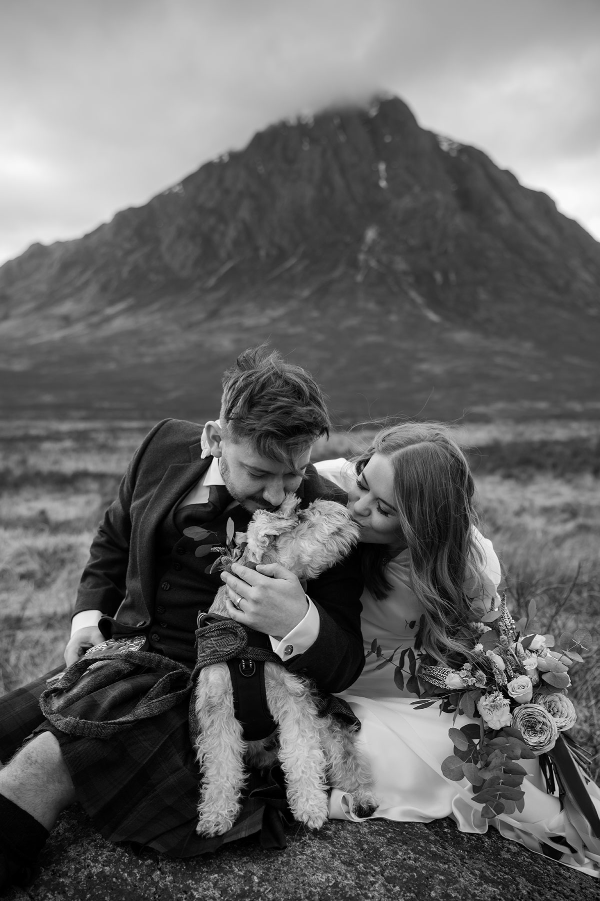 Black and white portrait of bride and groom cuddling their schnauzer dog on a rock in Glencoe after their Scottish elopement