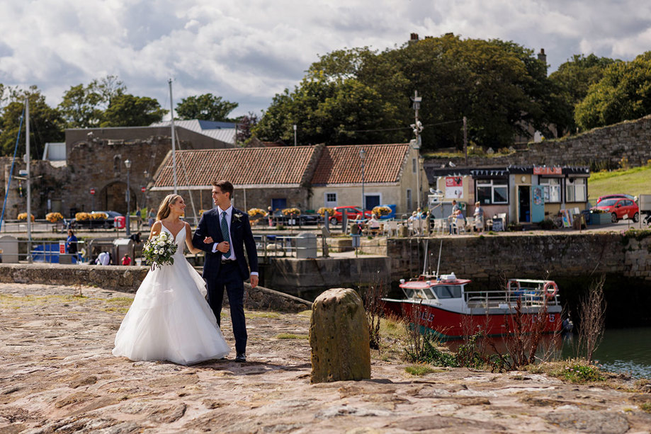 A bride and groom taking a leisurely stroll by the harbour on their St Andrews wedding day, soaking in the joy with quaint buildings and docked boats adding to the picturesque scene captured by Duke Photography