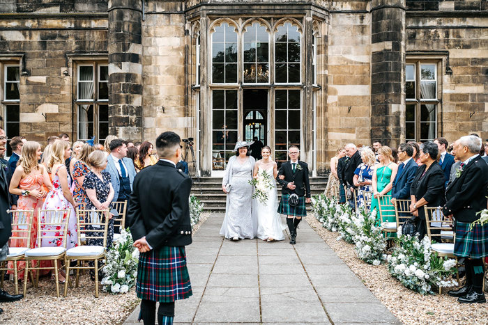 bride walks down the aisle with mum and dad towards groom wearing kilt for outdoor ceremony at mar hall
