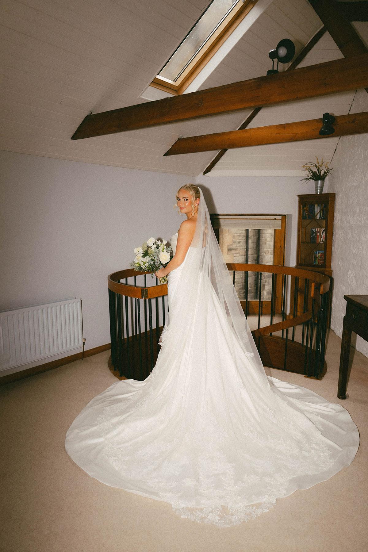 bride in wedding dress standing on staircase landing holding bouquet indoors